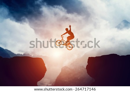 Man jumping on bmx bike over precipice in mountains at sunset. Raising hand showing hello gesture. Extreme sport, risk, cycling.