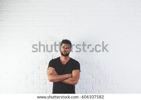 Portrait of handsome, thoughtful young man with folded amrs looking up on white brick background