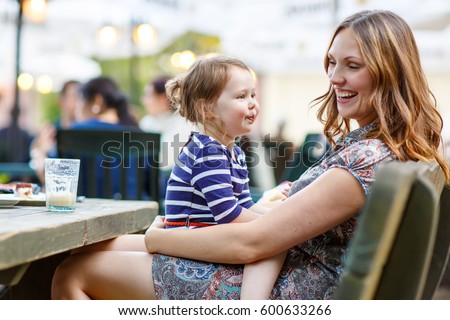 Young mother relaxing together with her little child, adorable toddler girl, in summer outdoors cafe waiting for coffe. Happy family hugging, beautiful woman and tiny cute kid.