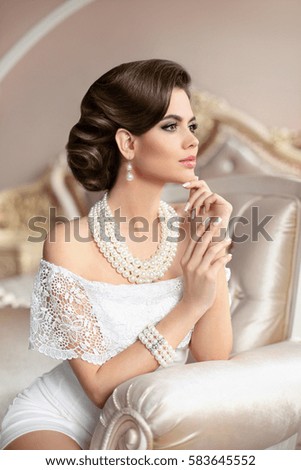 Retro woman portrait. Elegant brunette lady with fashion pearls jewelry set, french manicure nails, wavy hairstyle and makeup. Gorgeous model posing on luxury armchair in modern interior.
