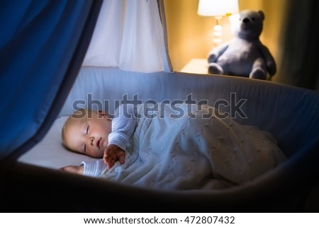 Adorable baby sleeping in blue bassinet with canopy at night. Little boy in pajamas taking a nap in dark room with crib, lamp and toy bear. Bed time for kids. Bedroom and nursery interior.
