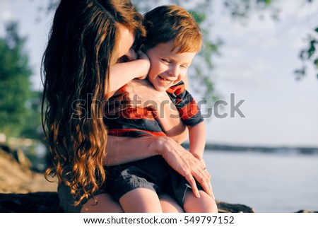 Young mother resting with his young son in the summer at sunset on the lake under a tree and hugs him. The child laughs and happy