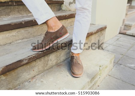 Brown perforated suede summer shoes with laces. Man in white trousers on the steps in the city. Men's summer fashion, stylish men's shoes.