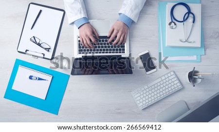 Doctor sitting at office desk and working on his laptop with medical equipment all around, top view