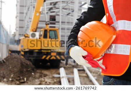 Construction safety concept, close-up worker wearing safety vest holding helmet with crane in the background