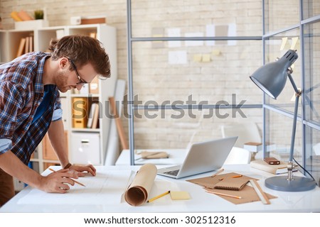 Modern architect looking at blueprint with sketch of construction in office