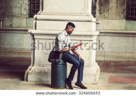 African American College Student studying in New York. Wearing gray shirt, jeans, cloth shoes, carrying shoulder leather bag, a black man sitting on metal pillar on street, relaxing, reading red book.