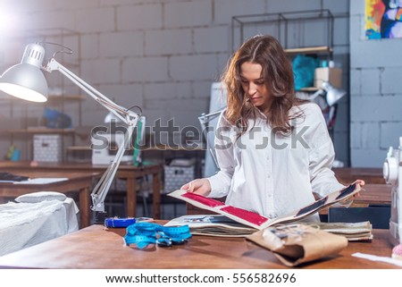 Young female dressmaker choosing material from catalogue in studio. Tailor looking through fabrics while standing in a sewing workshop