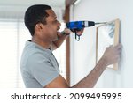 Small photo of Smiling black man builder drilling a hole in white wall and putting picture frame at home. Home renovation and decoration. Adult making DIY workshop. Concept for home diy, self service.