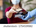 Small photo of Cashier hand holding credit card reader machine and wearing disposable gloves while client holding phone for NFC payment. Woman wearing face mask while paying with smartphone during Covid-19 pandemic.