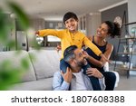 Small photo of Cheerful indian son sitting on father shoulder playing at home with african mother. Playful little boy enjoying spending time with parents at home. Flying child enjoying playing with his ethnic family