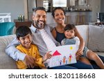 Small photo of Proud parents showing family painting of son and daughter sitting on sofa at home. Smiling african mother and middle eastern father with two children looking at camera. Brothers showing painting.