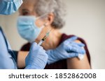 Small photo of Close up of general practitioner hand holding vaccine injection while wearing face protective mask during covid-19 pandemic. Young woman nurse with surgical mask giving injection to senior woman.