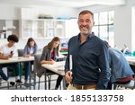 Small photo of Portrait of mature teacher looking at camera with copy space. Happy mid adult lecturer at classroom standing after giving lecture. Satisfied high school teacher smiling while his students studying.