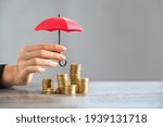 Small photo of Young woman hand holding small red umbrella over pile of coins on table. Close up of stack of coins with female hands holding umbrella for protection. Financial safety and investment concept.