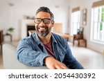 Small photo of Happy mature middle eastern man wearing eyeglasses sitting on couch. Portrait of indian man relaxing at home and looking away with big smile. Mid adult guy with specs thinking about his future.