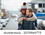 Small photo of Smiling man giving piggyback ride to woman in the city. Young multiethnic couple in cold clothes walking in street and having fun. Cheerful girlfriend with wool cap and boyfriend in sweater enjoying.