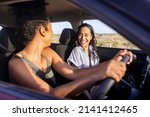 Small photo of Beautiful latin hispanic woman with friend enjoying and laughing in car while going on a road trip. Cheerful girl friends enjoying trip and drive in car at sunset. Black woman driving a car.