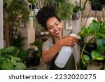 Small photo of Beautiful black florist taking care of plants while spraying it with water. African american owner working and spraying water plants in store. Happy and smiling florist watering plants in shop.