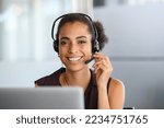 Small photo of Call center agent with headset working on support hotline in modern office. Young african american agent in conversation with customer over headset looking at camera. Portrait of black girl working.