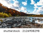Mountain river wild stone bank with rapids against colorful trees growing on hill slopes under blue sky with floating clouds in autumn