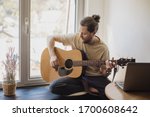 bearded man is sitting at home next to the window and playing the acoustic guitar
