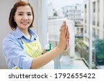 Portrait of pretty smiling Asian housewife wiping apartment window and looking at camera