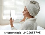 Young woman looking in mirror in her hand after morning shower while standing in bathroom and having skincare procedure