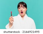 young pretty woman wearing bathrobe scared expression and holding a lipstick