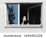 Man in medical protective face mask looking through window.Coronavirus pandemic covid-19.Man in corona quarantine looking out of window to the street.Man with face mask at home.During covid-19 quarant