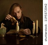 Small photo of long-haired man breaking away from reading an old book burns a dry leaf with a candle fire