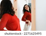 Small photo of Young beautiful self confident woman in red sweater looking in the mirror proud of herself