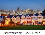 The Painted Ladies of San Francisco, California sit glowing amid the backdrop of a sunset and skyscrapers.