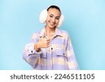 Small photo of Young Arab woman wearing winter muffs isolated on blue background with thumbs up because something good has happened