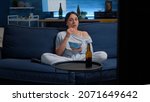 Small photo of Scared young woman watching horror film on TV eating popcorn closing eyes frightened sitting on sofa at night in modern apartment. Awestruck and wide-eyed lady reacting to astonishing moment on tv