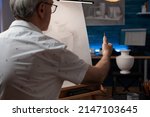 Small photo of Focus on hand of creative senior artist looking at plaster vase measuring proportions using pencil doing creative pencil drawing. Elderly man in the evening sketching scale model in home art studio.