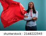 Small photo of Brave and happy looking young adult superhero woman wearing red hero cape while smiling at camera. Justice defender with superpowers and mighty posture standing with arms crossed on blue background.