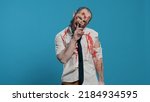 Small photo of Creepy dead walking corpse making shush secrecy hand gesture on blue background. Dangerous walking dead corpse with deep and bloody wounds being confidential while smirking at camera.
