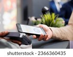 Small photo of Female hand paying in clothing store with smartphone, tapping phone on contactless card reader while standing at cash register, close up. Customer using mobile phone as payment method in shopping mall