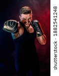 Small photo of Bearded tattooed sportsman muay thai boxer in black undershirt and boxing gloves fighting on dark background with smoke. Sport concept.