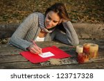 woman thoughtfully smiling, leaning on the arm, writing word LOVE on white and red paper on wooden table with candles