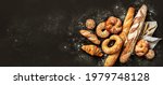 Bakery - various kinds of breadstuff. Bread rolls, baguette, bagel, sweet bun and croissant captured from above (top view, flat lay). Black background, free copy space. Horizontal banner layout.