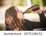 Small photo of Girl chug beer from bottle on wooden bench with table in forest park outdoor. Express drinking and fast get drunk lifestyle concept. Real life scene