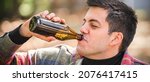 Small photo of Man chug beer from bottle on wooden bench with table in forest park outdoor. Express drinking and fast get drunk lifestyle concept. Real life scene