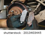 Small photo of The car mechanic shows for comparison a worn brake shoe and a new one against the background of an automobile brake disc with signs of corrosion