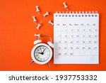 Small photo of white calendar page , alarm clock and thumbtacks on grunge orange paper background