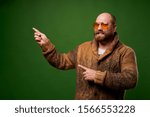 Small photo of Bald male in sweater pointing his hand to empty place against green background
