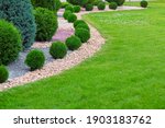 Landscape bed of garden with wave ornamental growth cypress bushes gravel mulch by color rock way on a day spring park with green lawn meadow, nobody.