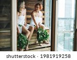 Young women friends in white towel sitting on wooden shelf in Finnish sauna Russian bath with rowan brooms in spa hotel in front of panoramic window, treat yourself