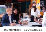 Small photo of positive female busboy in mask serving order to friends in modern restaurante